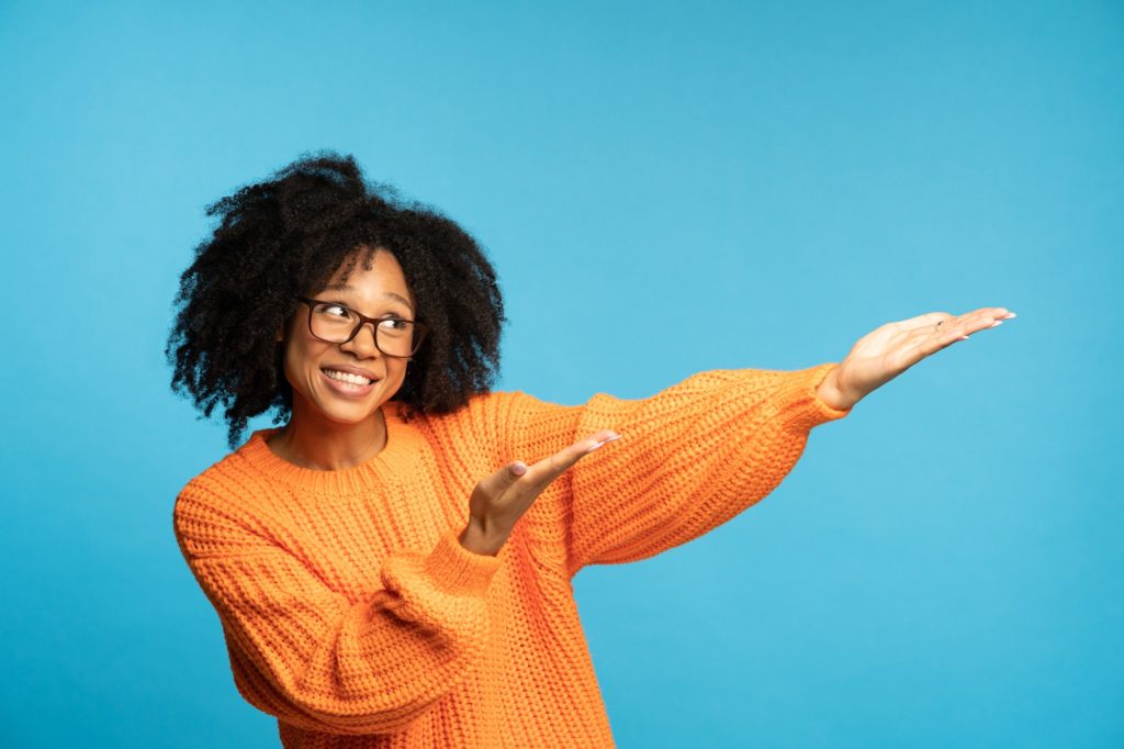 Biracial woman with curly hair in orange jumper points away with hands, showing at blank copy space
